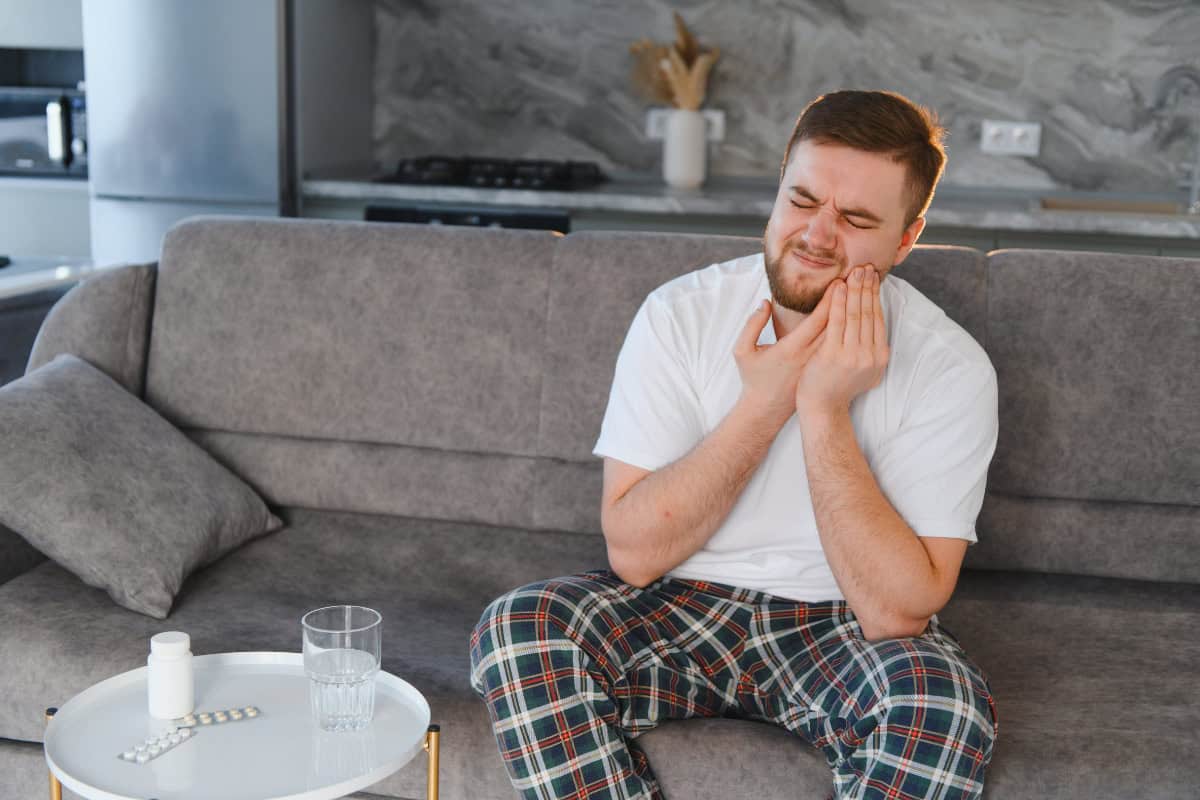 Man on a gray sofa holding his jaw in pain; pills and a glass on a nearby white table.