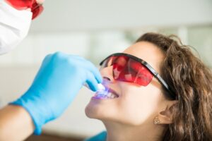 Dentist wearing a mask and blue gloves uses a blue light device on a patient’s teeth, who wears red protective glasses.