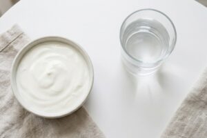 Bowl of creamy white yogurt on a beige napkin next to a glass of water on a white table.