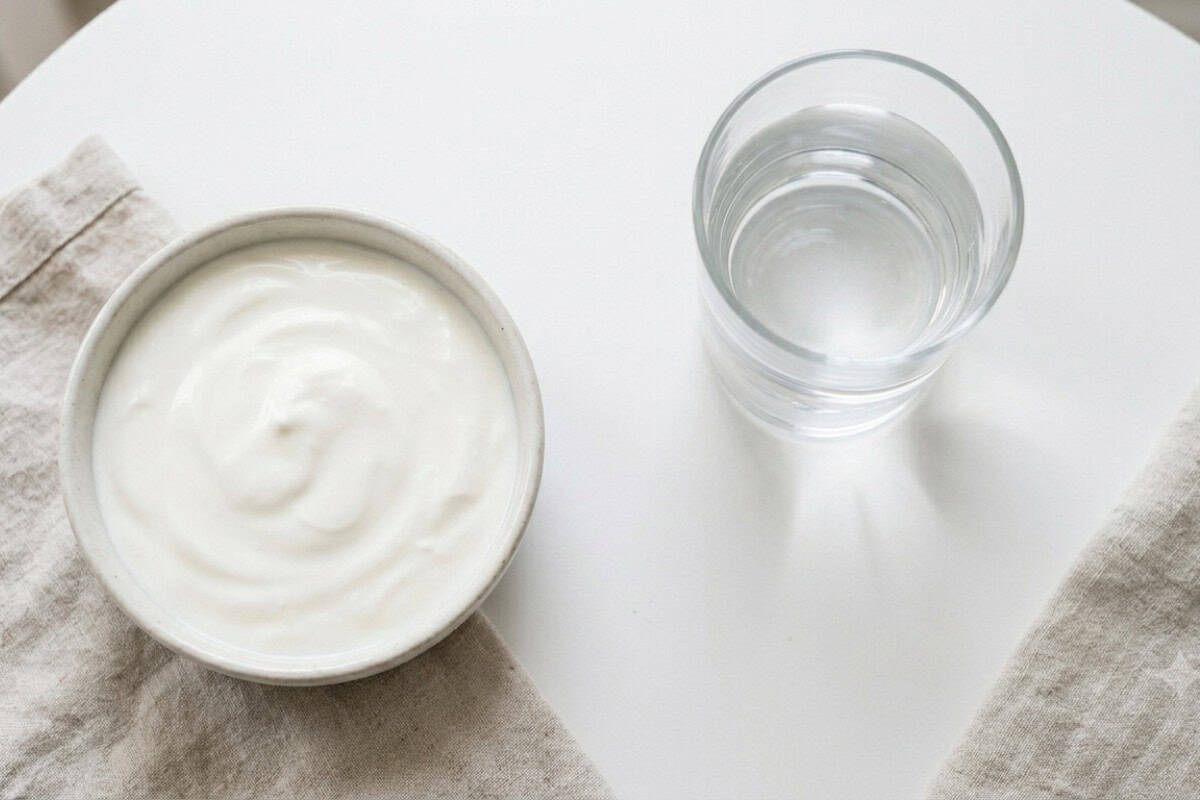 Bowl of creamy white yogurt on a beige napkin next to a glass of water on a white table.
