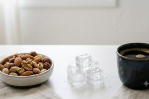 what-foods-to-avoid-with-veneers-why-it-matters - Magnolia Family Dental Care | Tulsa Dentist | Tulsa, OK Bowl of mixed nuts on a white table with ice cubes in the center and a dark coffee cup on the right.