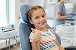 Young girl in a dental chair smiling and giving a thumbs up with a clinician in the background.