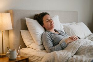 Woman resting in bed with eyes closed, smiling softly, hands folded on chest; bedside table with lamp and mug nearby