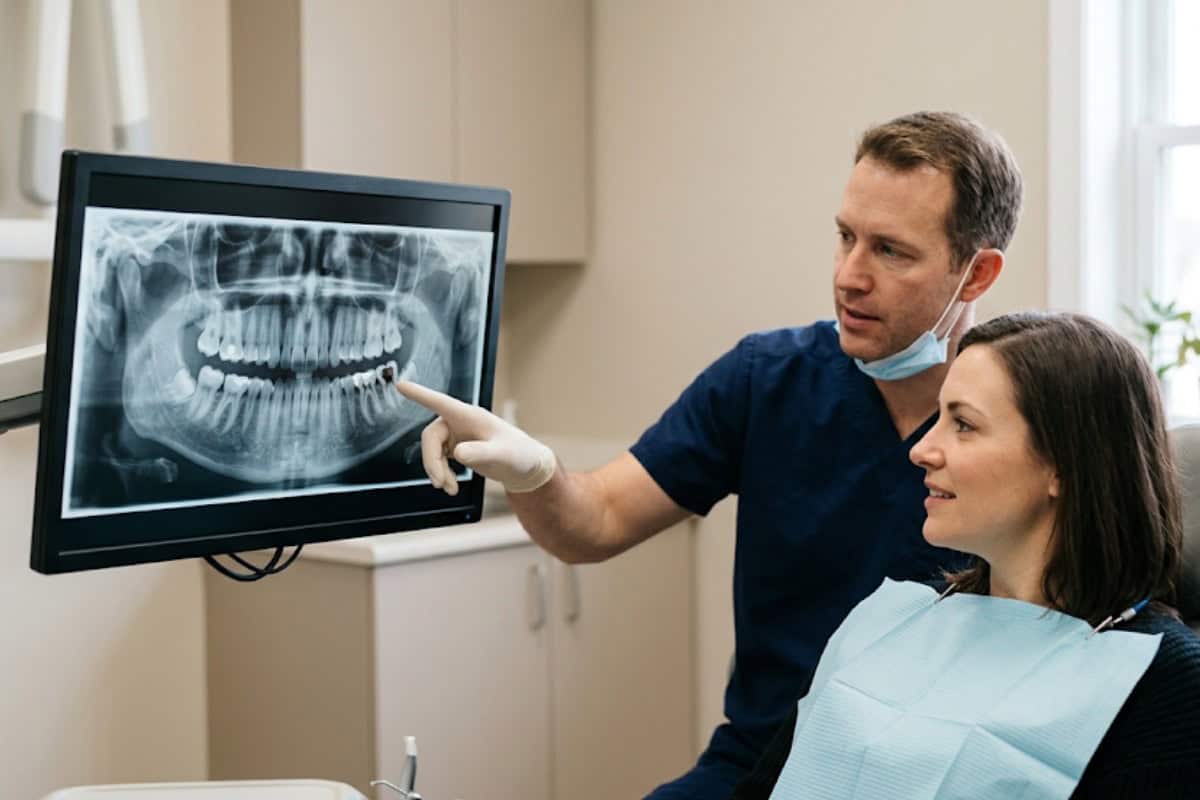 Dentist in blue scrubs points to a dental X-ray of teeth on a monitor while a patient sits in the chair.